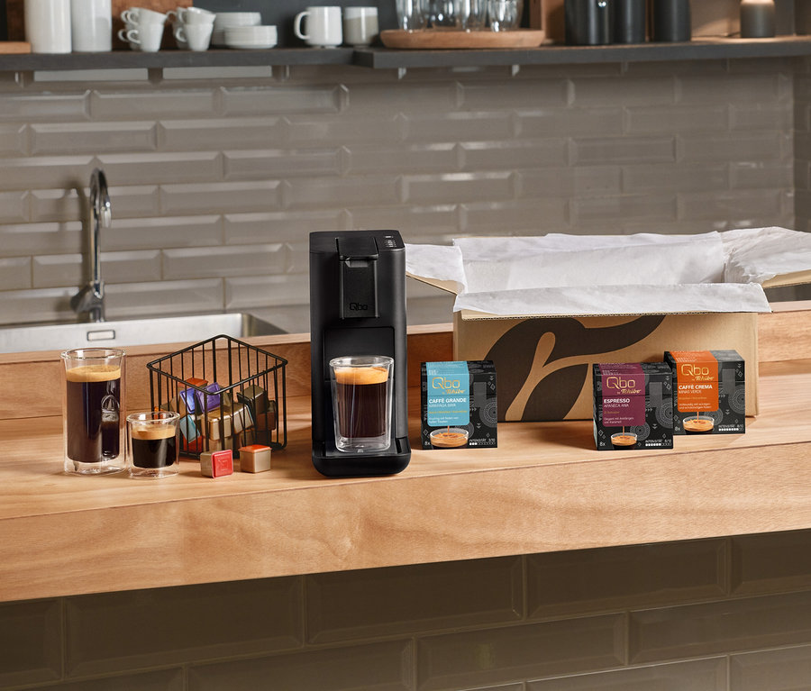 Eine schwarze Qbo Iconic, Dark Chrome Kaffeemaschine auf einer Holztheke, mit einem Glas Kaffee darunter. Daneben stehen ein großes und ein kleines Glas Kaffee, ein Korb mit Kapseln und drei Packungen Qbo Kapseln, darunter Qbo Caffè Grande KIRINYAGA BAYA und Qbo Espresso APANECA ANA. Im Hintergrund ist ein offener Karton zu sehen.