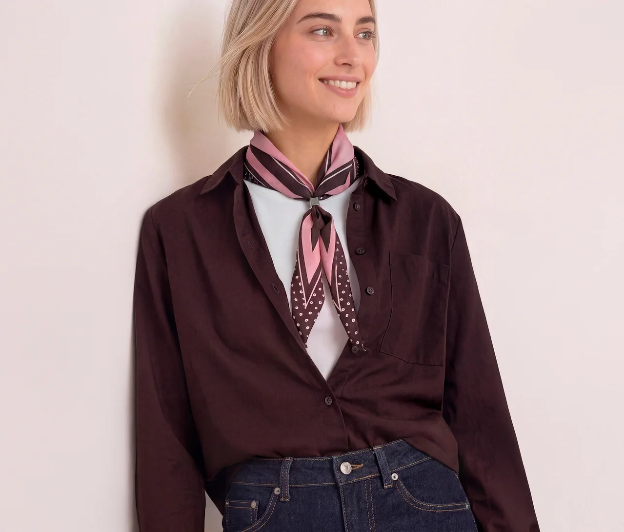 Eine lächelnde Frau mit kurzen blonden Haaren trägt ein Street One Bandana in Rosa und Dunkelbraun mit Streifen und Punkten, ein offenes dunkelbraunes Hemd und Jeans.