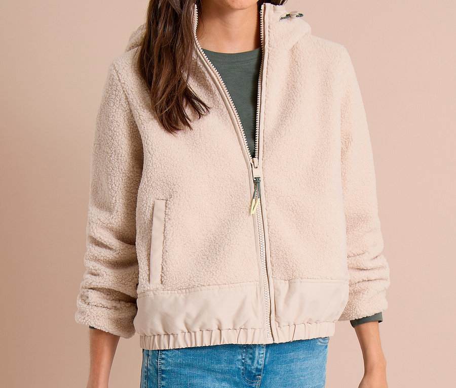 Lächelnde Frau in beiger CECIL Fleece-Reißverschlussjacke mit flauschigem Oberteil und glattem Unterteil, grünem Oberteil und blauer Jeans, die einen schwarzen Gegenstand hält.