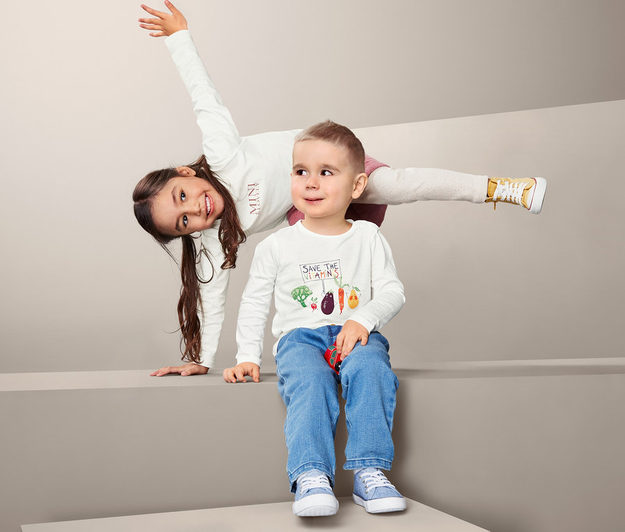 Zwei Kinder posieren vor grauem Hintergrund. Mädchen im Langarmshirt steht auf ihren Händen, Junge sitzt in Jeans.