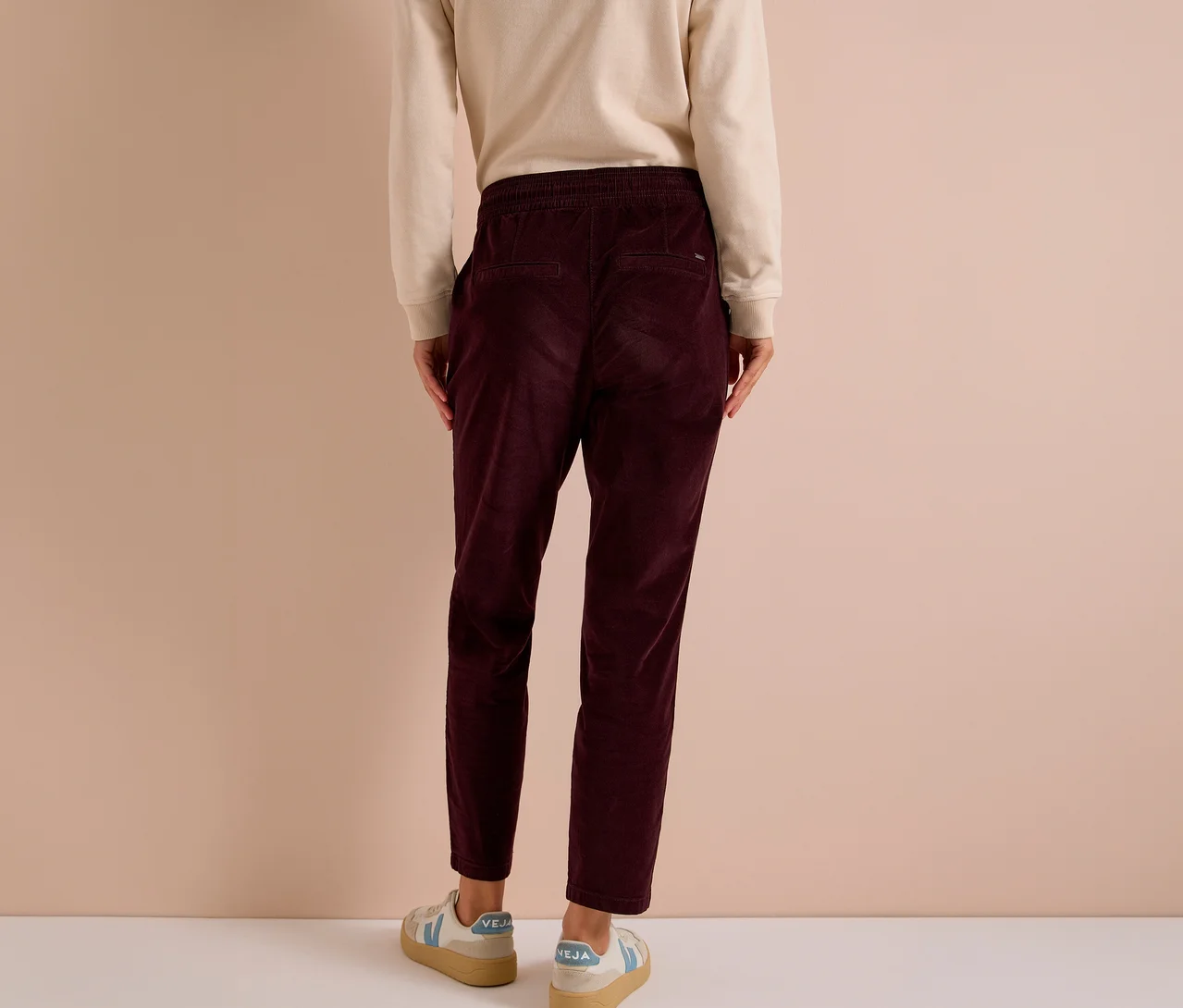 Person von hinten in einem hellen Langarmshirt, einer dunkelroten CECIL Cordhose mit Gummibund und hellen Sneakern mit blauen Akzenten.