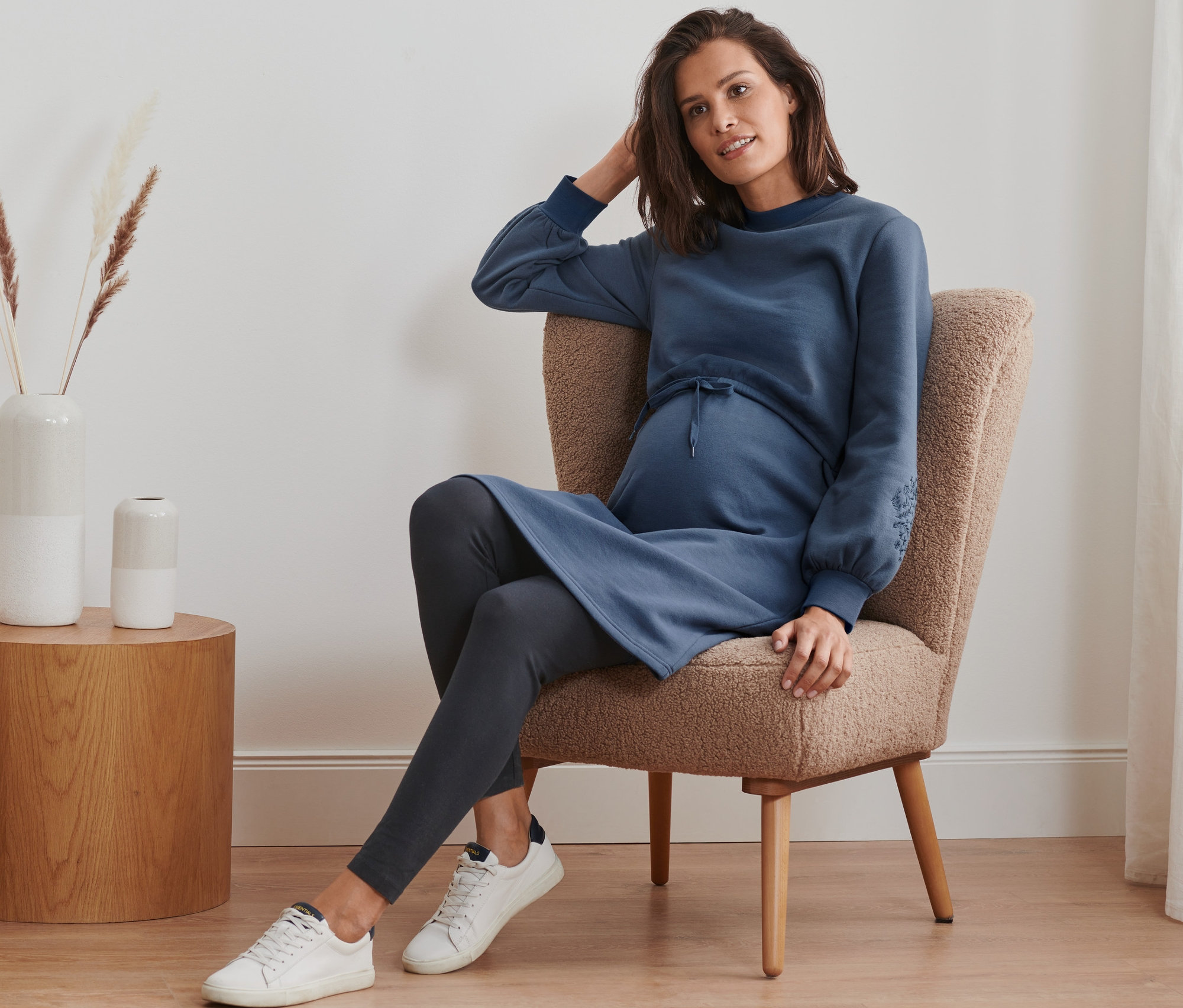 Schwangere Frau sitzt im Sessel und trägt Still-/Umstands-Sweatkleid, Leggings und Sneakers.