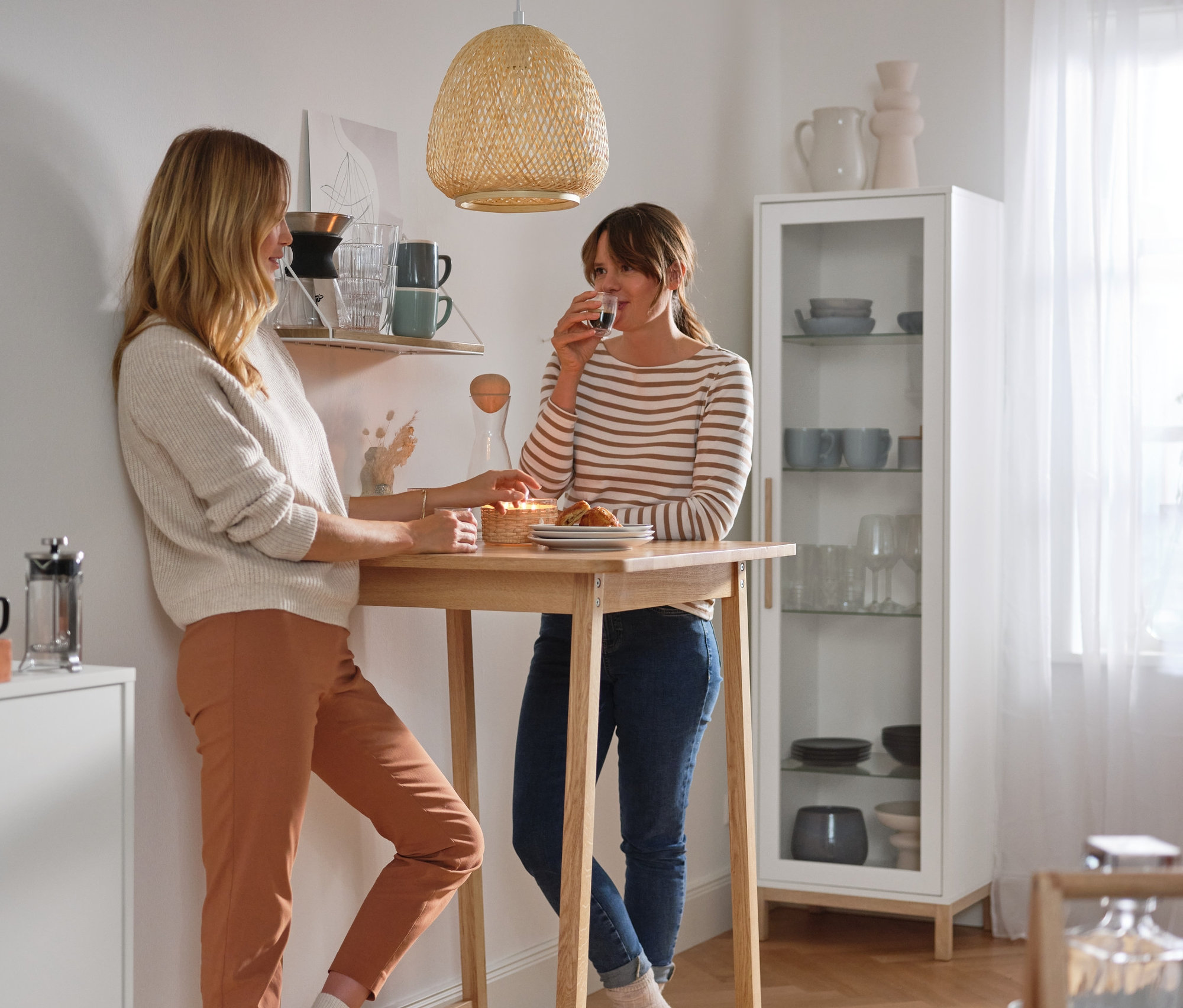 Zwei Frauen stehen an einem Bartisch und unterhalten sich. Im Hintergrund steht eine hohe Vitrine »Eklund« mit Glastür.