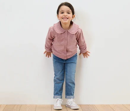 Lächelndes Mädchen steht vor einer weißen Wand und trägt Jeans und eine Kinder-Sweatjacke mit Volant.