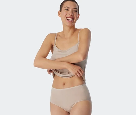 Lächelnde Frau mit kurzen Haaren zieht ihr sandfarbenes Top hoch und trägt sandfarbene SCHIESSER Midislips mit Modal.