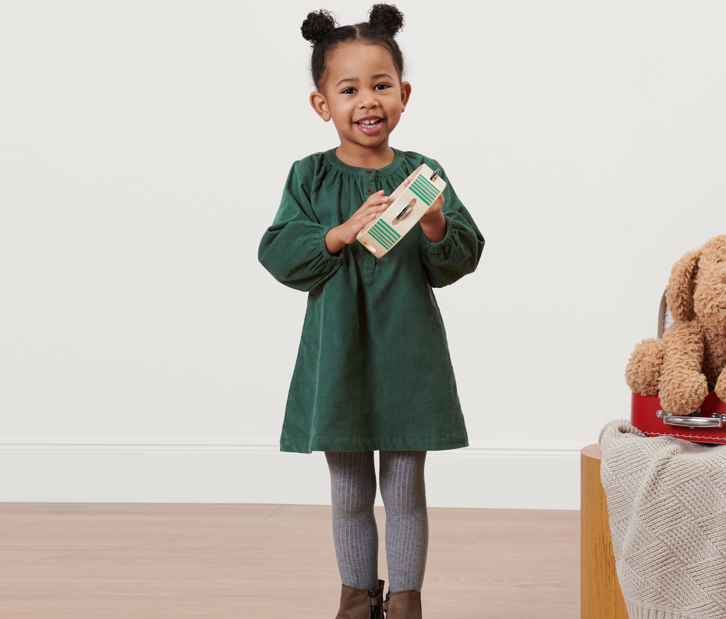 Lächelndes kleines Mädchen hält ein Holzspielzeug und trägt ein grünes Kleinkind-Cordkleid, graue Leggings und braune Stiefel.