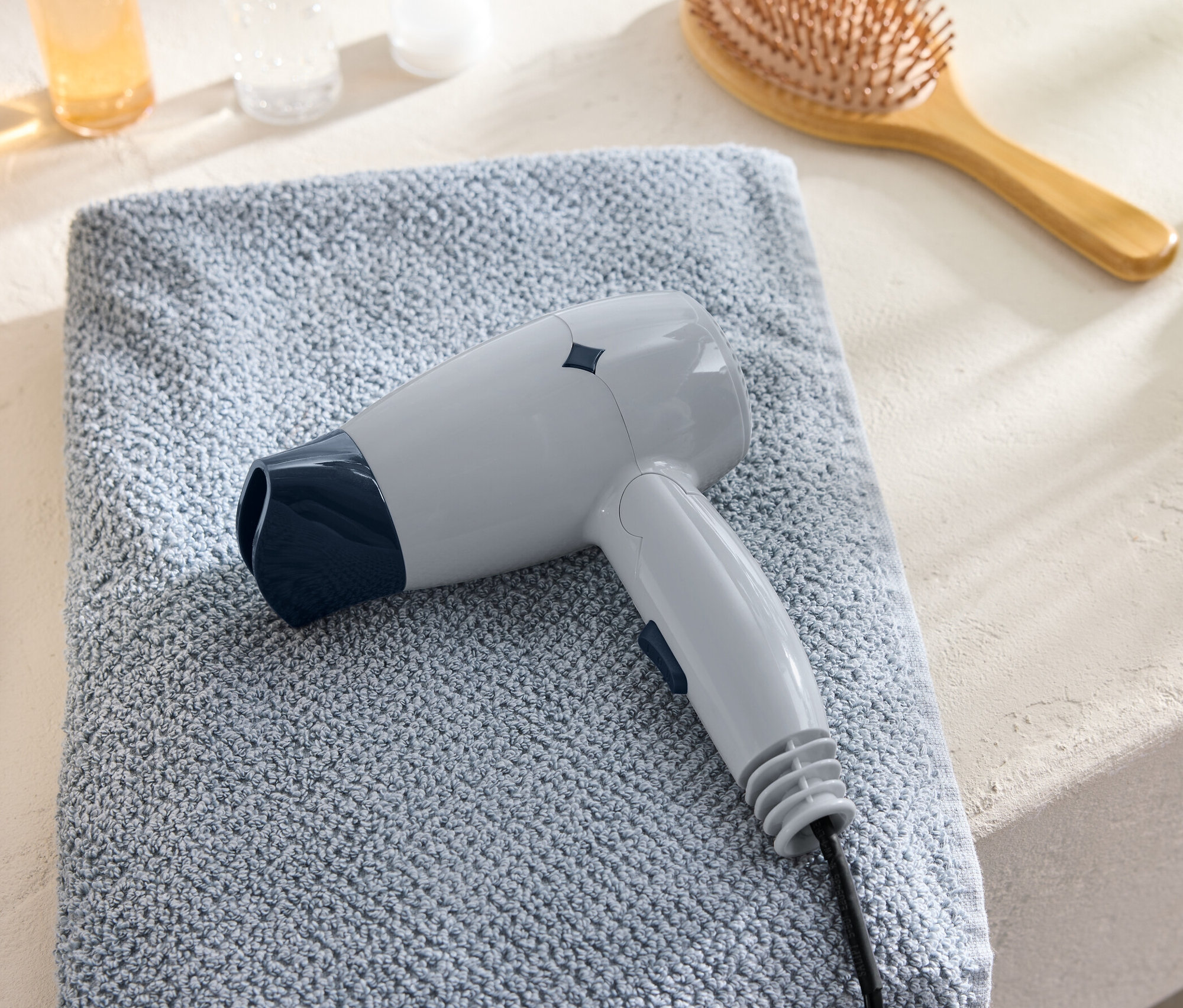 Ein grauer Reise-Haartrockner liegt auf einem blauen Handtuch neben einer Bürste.