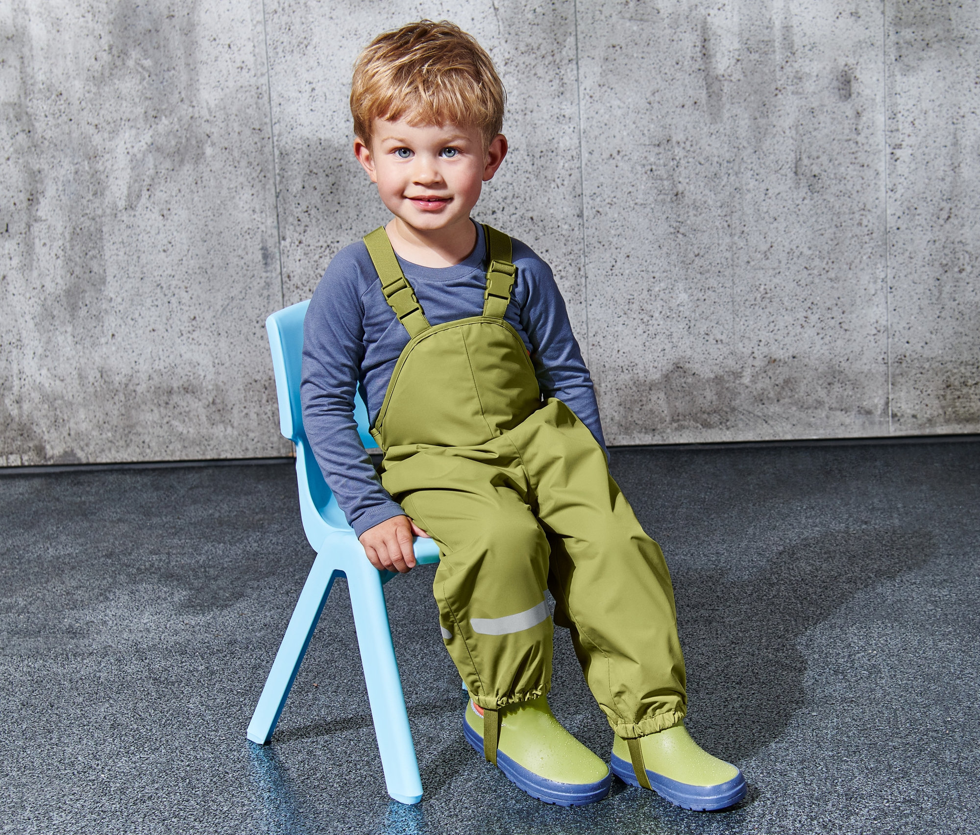 Ein Junge sitzt auf einem blauen Stuhl und trägt eine grüne Kleinkinder-Regenhose.