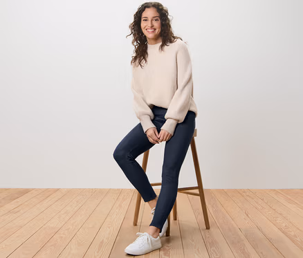 Frau sitzt auf einem Hocker und trägt eine gefütterte Leggings in Jeansoptik und einen Pullover.