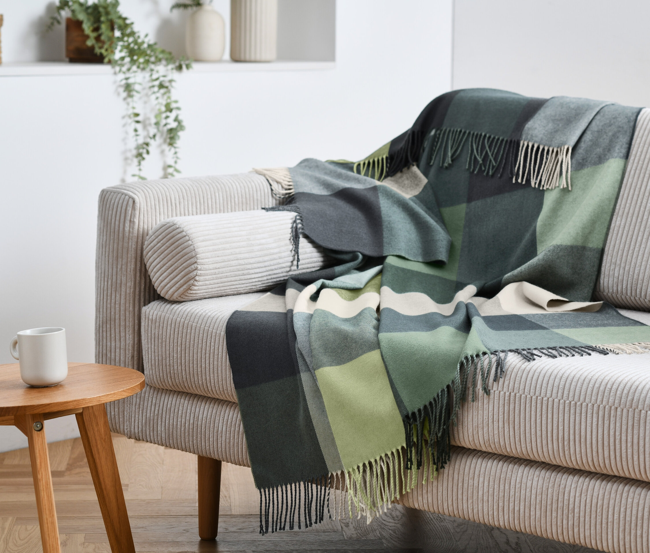 Ein karierter grüner Plaid mit Fransen liegt auf einem beigen Sofa. Auf einem Holztisch steht eine weiße Tasse.
