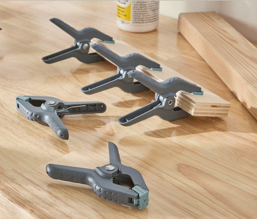 Auf einer Holzoberfläche liegen ein Bohrer und fünf graue Klemmzwingen.