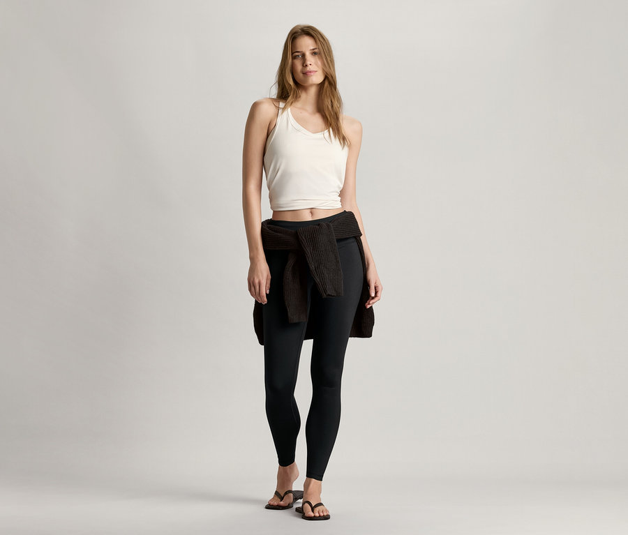 Frau steht und trägt ein weißes Seamless-Sport-Bustier, eine schwarze Leggings und eine Strickjacke um die Hüften.