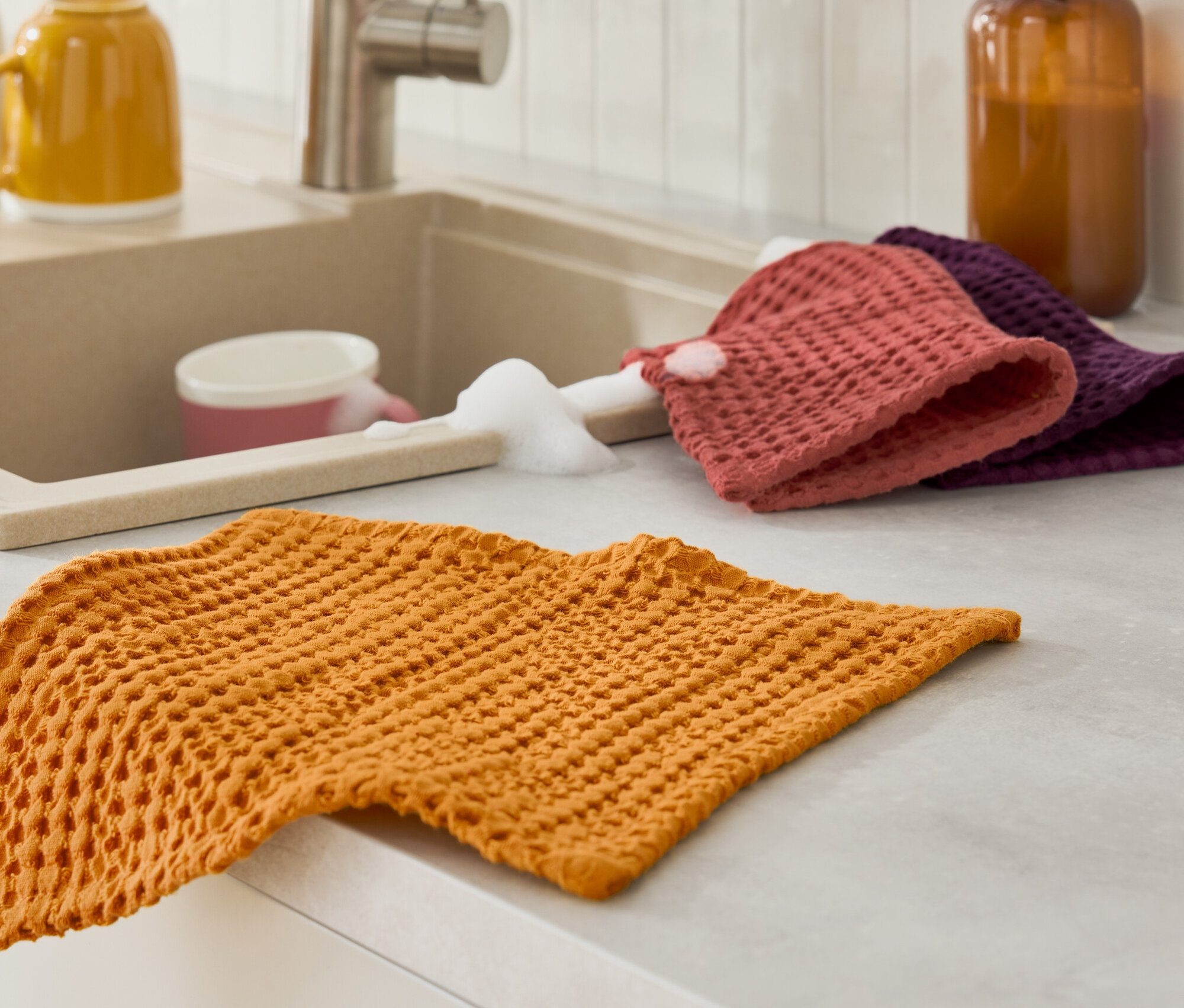 Ein orangefarbenes Geschirrtuch liegt auf der Arbeitsfläche neben dem Spülbecken mit Schwamm und Spülmittel.