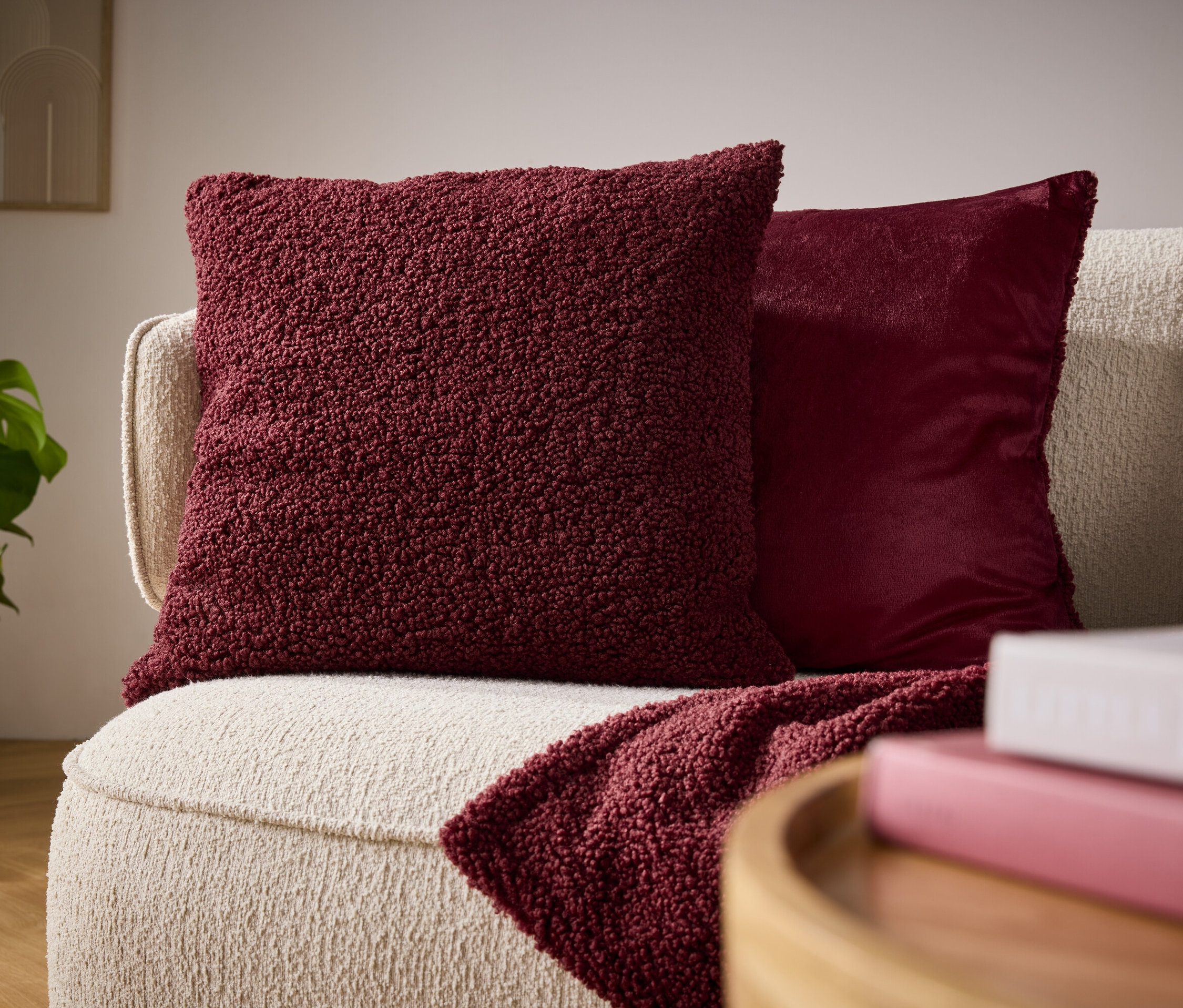 Detail eines beigen Sofas mit einem bordeauxroten Plüschkissen, einem glänzenden Kissen und einer drapierten Decke.