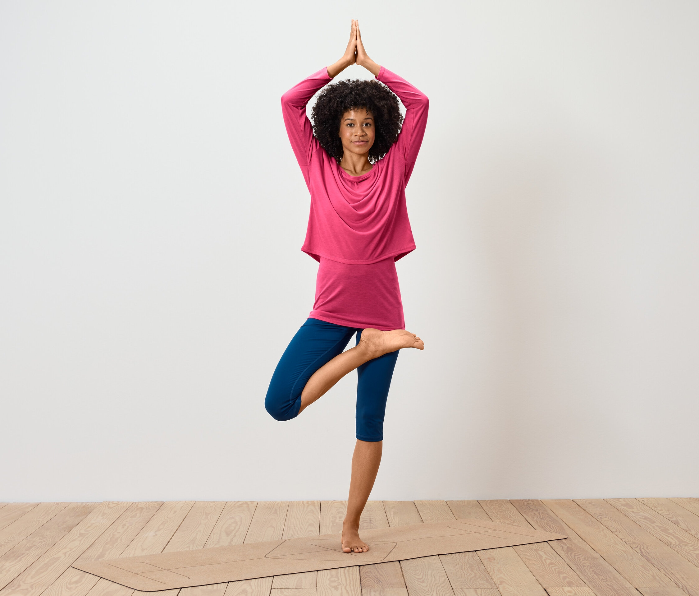 Frau mit Afro-Frisur steht in der Baum-Pose auf einer Yogamatte, trägt ein pinkfarbenes Langarmshirt und blaue Leggings.