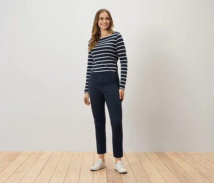 Eine Frau steht und trägt ein langärmliges Streifenshirt, dunkelblaue Hosen und weiße Turnschuhe.