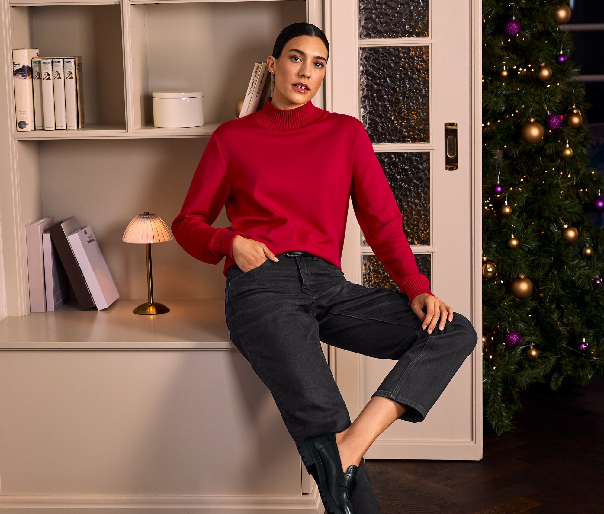 Eine Frau sitzt neben einem Bücherregal und einem Weihnachtsbaum und trägt einen roten Sweatshirt aus Modal-Mix, eine Modern Jeans – Fit »Ava«, einen schwarzen Ledergürtel und Leder-Loafer.