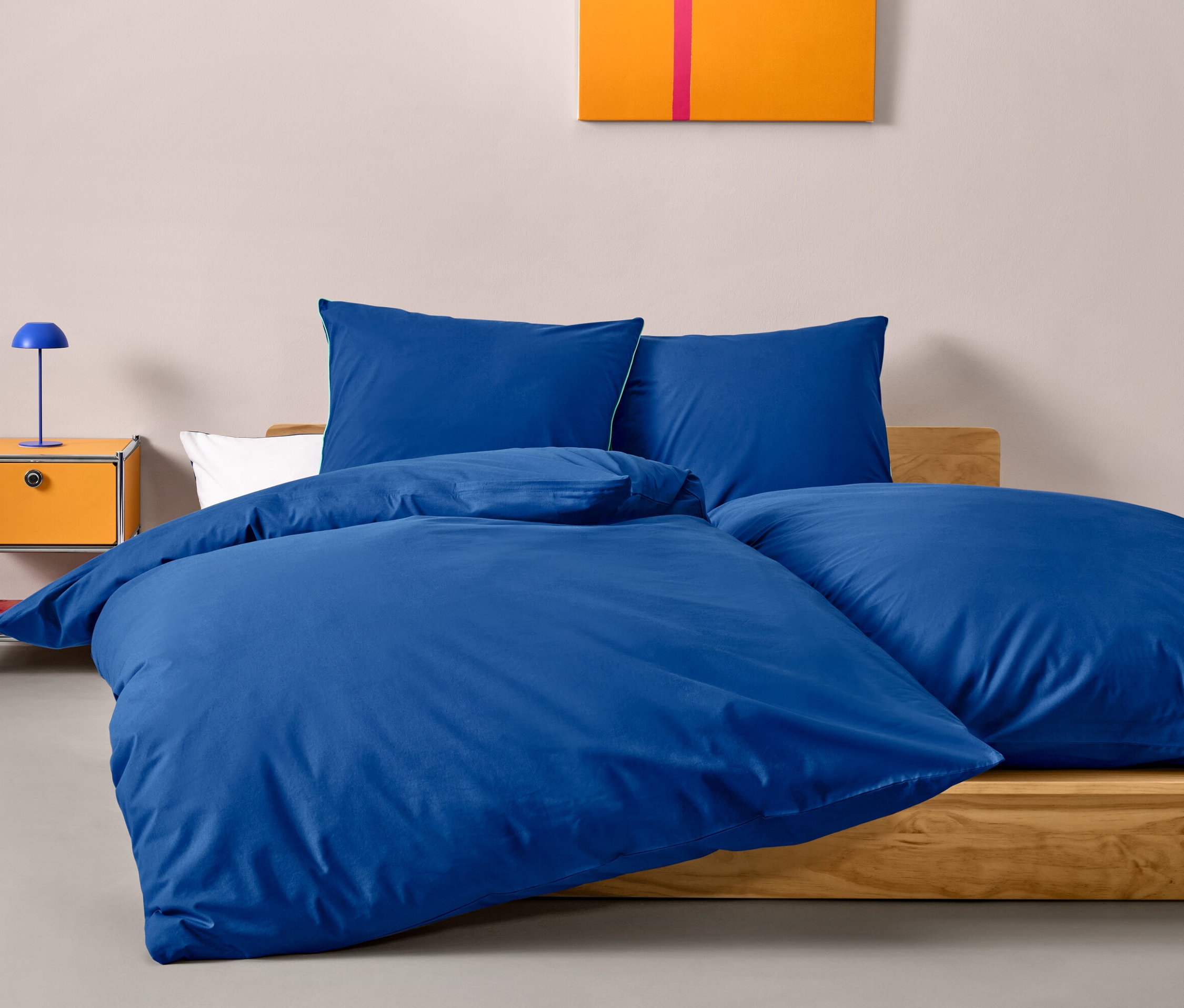 Blaue Renforcé-Bettwäsche in Normalgröße auf einem Bett, neben einem orangefarbenen Nachttisch mit blauer Lampe.