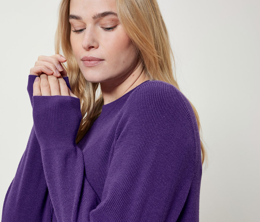 Blonde Frau in einem violetten Pullover hält ihre Hände an ihr Gesicht.