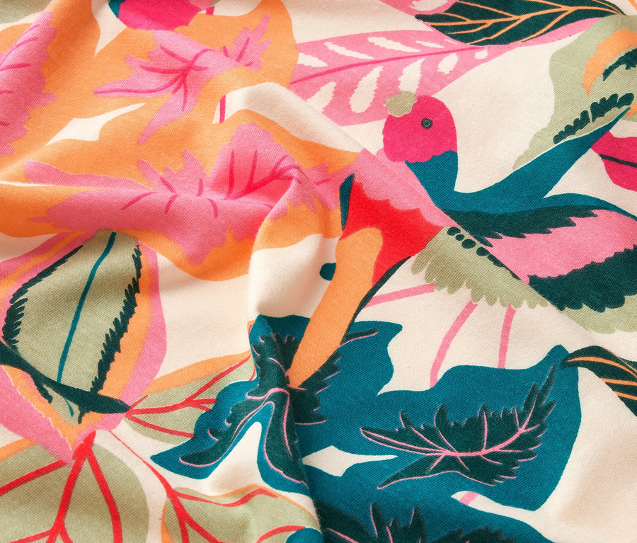 Nahaufnahme von Stoff mit Blatt- und Vogelmuster in Pink, Orange, Grün und Blau auf beigem Grund.