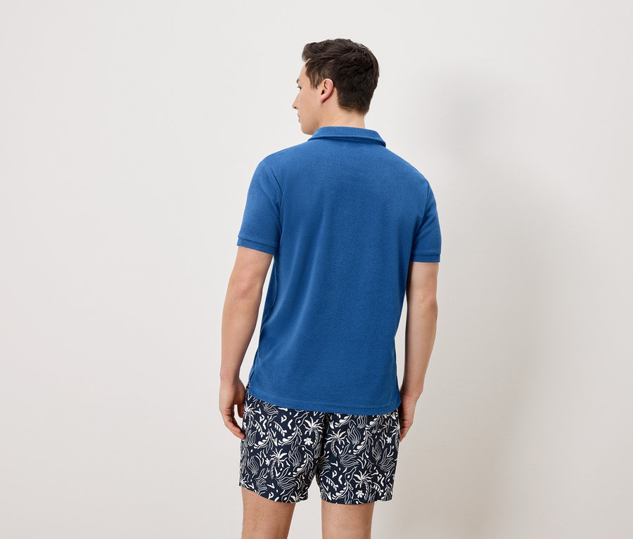 Ein Mann von hinten, der ein blaues Poloshirt und blau-weiße Badeshorts trägt.