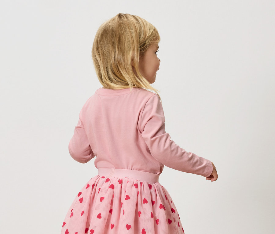 Kleines blondes Mädchen trägt ein rosa Langarmshirt und einen rosa Tüllrock mit roten Herzen.