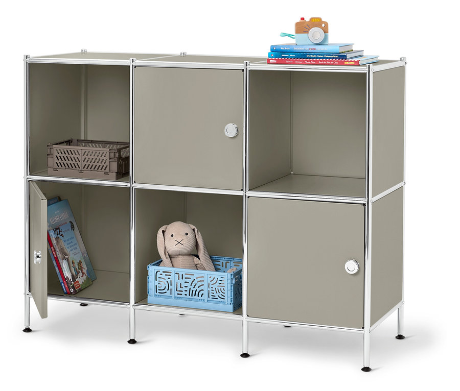 Ein graues modulares Regal mit sechs Fächern, davon drei mit Türen, gefüllt mit einer Spielzeugkamera, Büchern, einem Korb, einem Stofftierhasen und einem Kinderbuch.