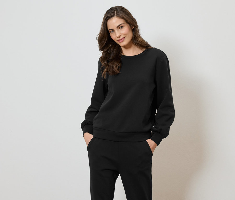 Frau trägt ein schwarzes Sweatshirt und eine Hose und posiert vor einem weißen Hintergrund.