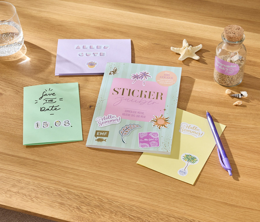 Auf einem Tisch liegen ein Glas Wasser, Karten mit Aufschriften, ein Stickerbuch, ein Stift, ein Seestern und eine Flasche mit Sand.