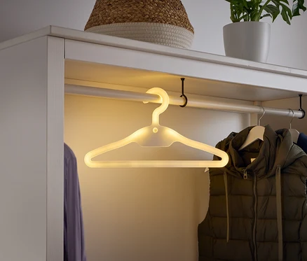 In einem weißen Kleiderschrank hängt ein leuchtender Kleiderbügel an einer Stange, neben einer Weste und einem Hemd.