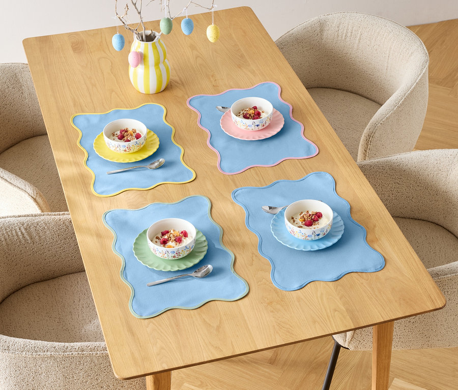 Ein Esstisch ist mit vier Gedecken mit Müslischalen und farbigen Platzsets dekoriert, umgeben von vier Stühlen.