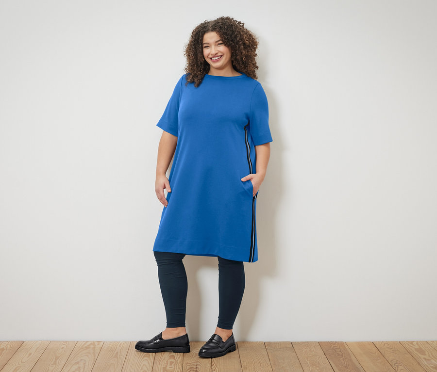 Frau mit lockigem Haar trägt ein blaues Kleid mit Reissverschlussdetail und Leggings, posiert vor einer Wand.