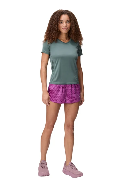 Frau steht mit grünem T-Shirt, pinken Shorts und pinken Schuhen vor weißem Hintergrund.