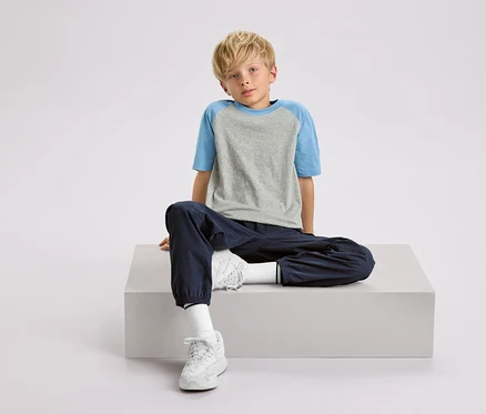 Ein blonder Junge sitzt auf einem weißen Würfel, trägt ein grau-blaues T-Shirt, marineblaue Hosen und weiße Turnschuhe.