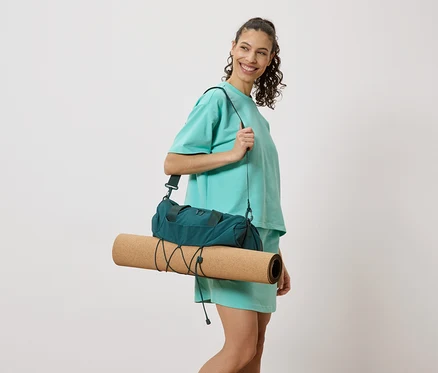 Eine Frau im türkisfarbenen Outfit mit einer dunkelgrünen Sporttasche und einer zusammengerollten Yogamatte über der Schulter, lächelnd.