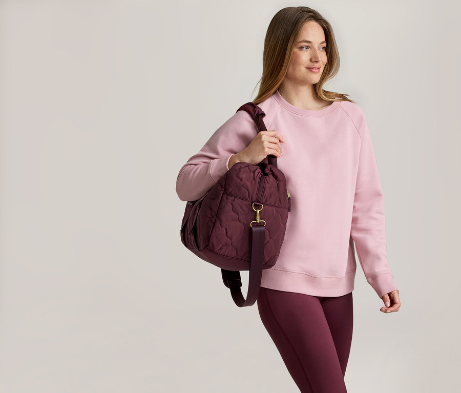Frau mit rosa Pullover und bordeauxfarbener Leggings, die eine gesteppte, bordeauxfarbene Tasche über der Schulter trägt.