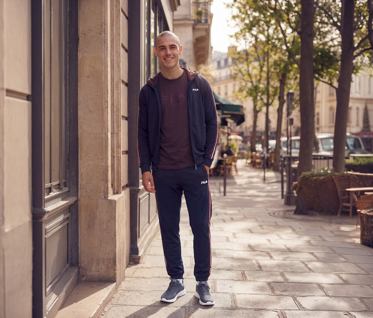 Lächelnder Mann in einem marineblauen Trainingsanzug mit bordeauxroten Streifen, einem bordeauxroten T-Shirt und grauen Sneakers, der auf einem Bürgersteig steht.