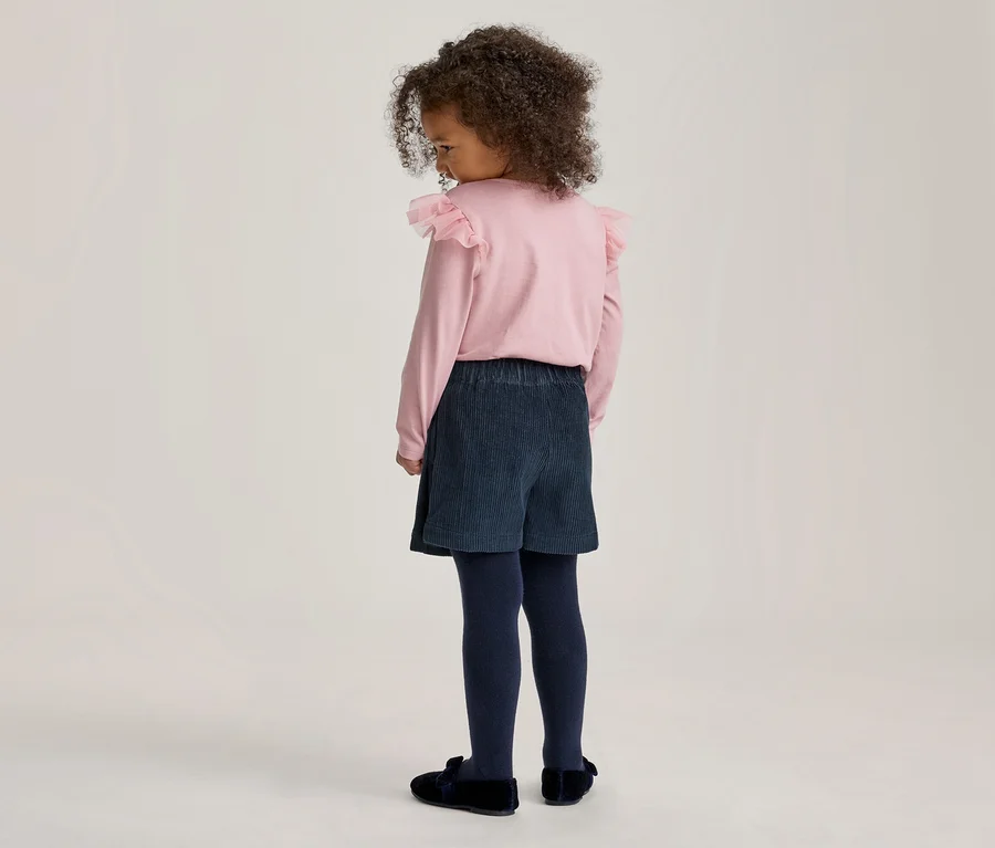 Kleines Kind mit lockigem Haar von hinten, trägt ein rosa Langarmshirt mit Rüschen an den Schultern, dunkelblaue Cord-Shorts, dunkelblaue Strumpfhosen und dunkelblaue Slipper.