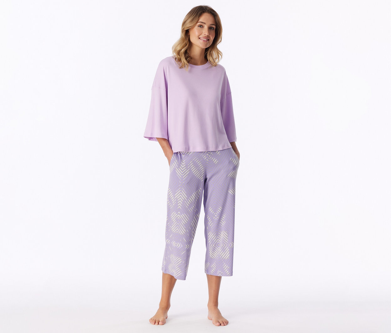 Lachende Frau steht barfuß, trägt ein hellviolettes Kurzarmshirt und hellviolette Caprihose mit weißem geometrischem Muster, Hände in den Taschen.