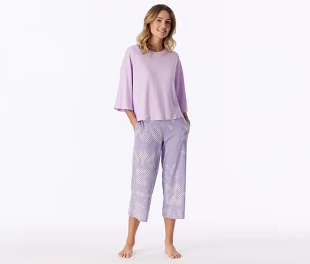 Lachende Frau steht barfuß, trägt ein hellviolettes Kurzarmshirt und hellviolette Caprihose mit weißem geometrischem Muster, Hände in den Taschen.