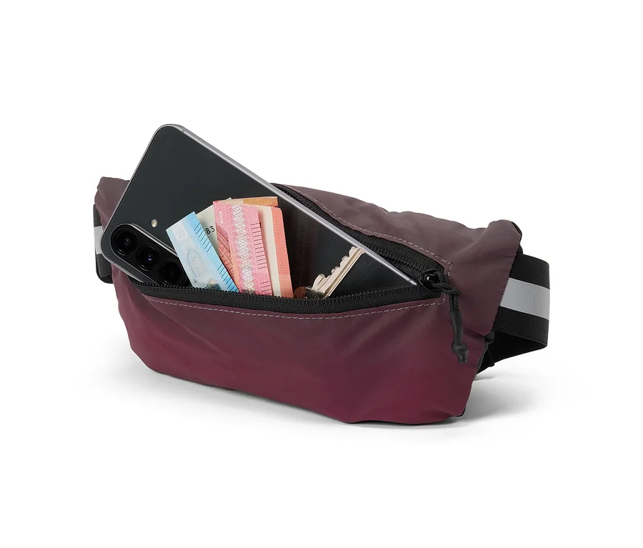 Bordeauxrote Bauchtasche mit geöffnetem Reißverschluss, in der ein schwarzes Smartphone, Geldscheine und ein Schlüssel stecken, auf weißem Hintergrund.