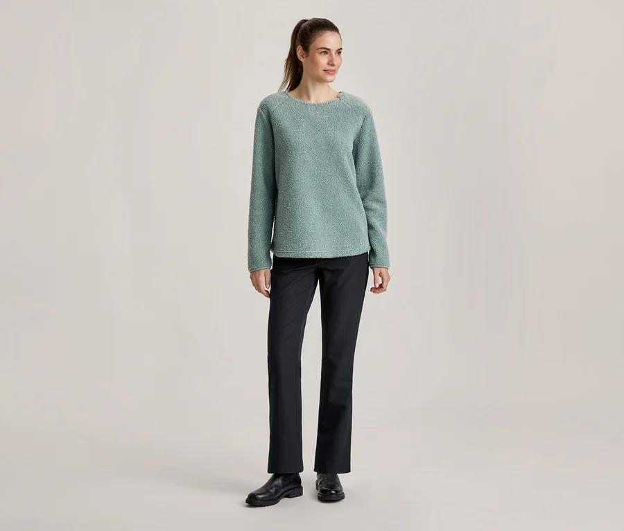 Stehende Frau in einem hellgrünen, strukturierten Pullover, schwarzen Hosen und schwarzen Schuhen, mit Pferdeschwanz.