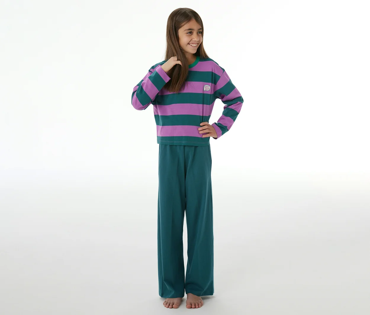Lächelndes Mädchen in einem SCHIESSER Kinder-Pyjama mit lila-grün gestreiftem Langarmshirt und petrolfarbener Hose, barfuß.