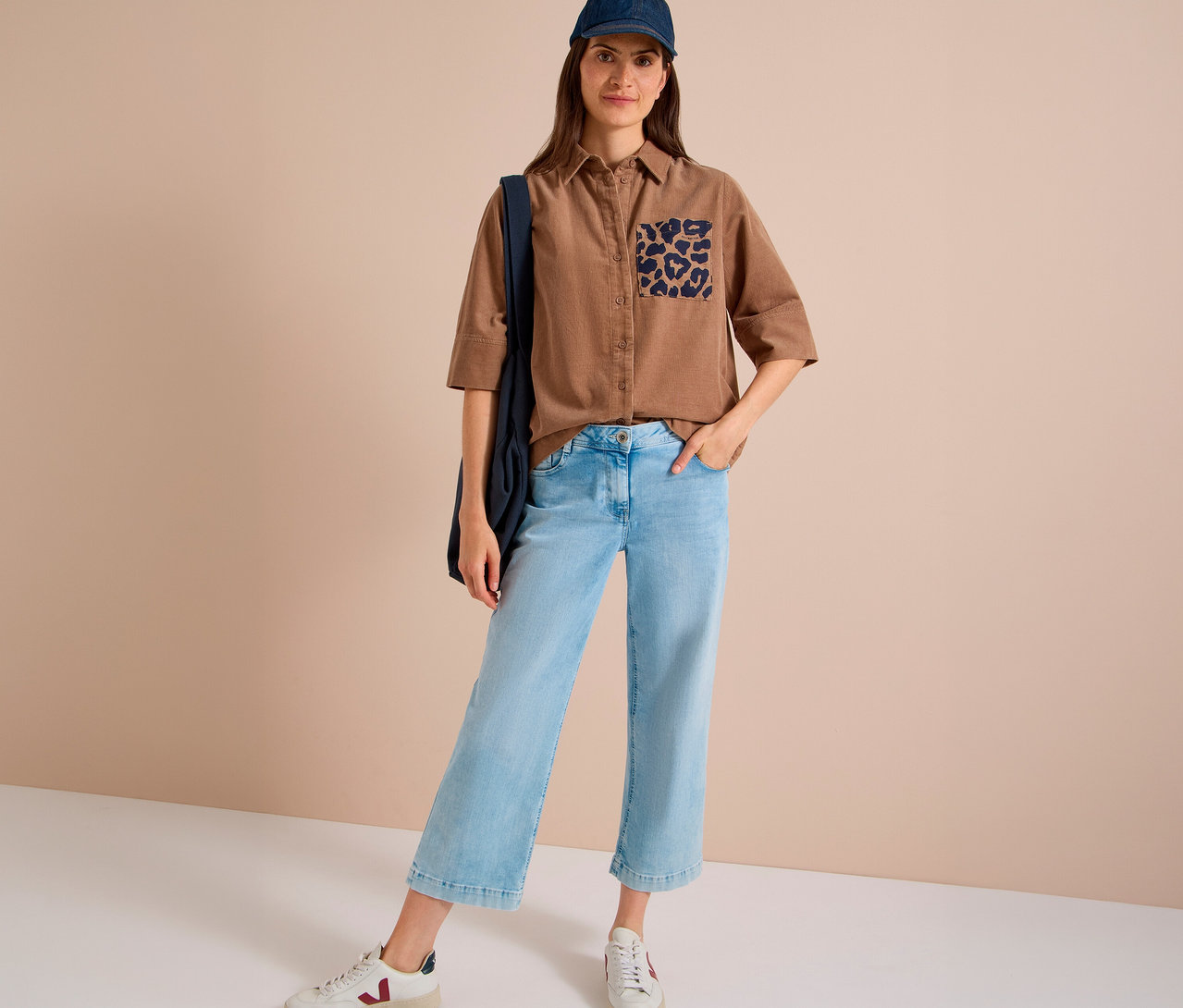 Stehende Frau in einer braunen CECIL Hemdbluse mit Leopardenmuster-Tasche, hellblauen Cropped-Jeans, blauer Baseballkappe und weißen Sneakern, eine dunkelblaue Tasche über der Schulter tragend.