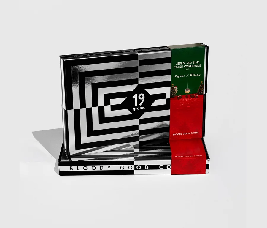 Teilweise geöffneter 19grams Adventskalender für Kaffee mit 24x 50 g ganzen Bohnen. Die Box ist schwarz-weiß gestreift, rot und grün, mit '19grams x Tchibo'.