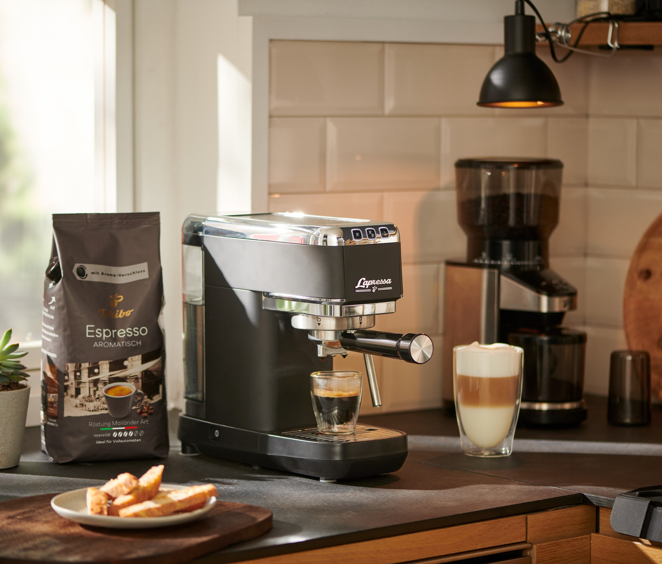 Espresso läuft aus einer »Lapressa« Siebträgermaschine, schwarz, in ein Glas. Im Hintergrund stehen eine Kaffeemühle und eine Packung Kaffee.