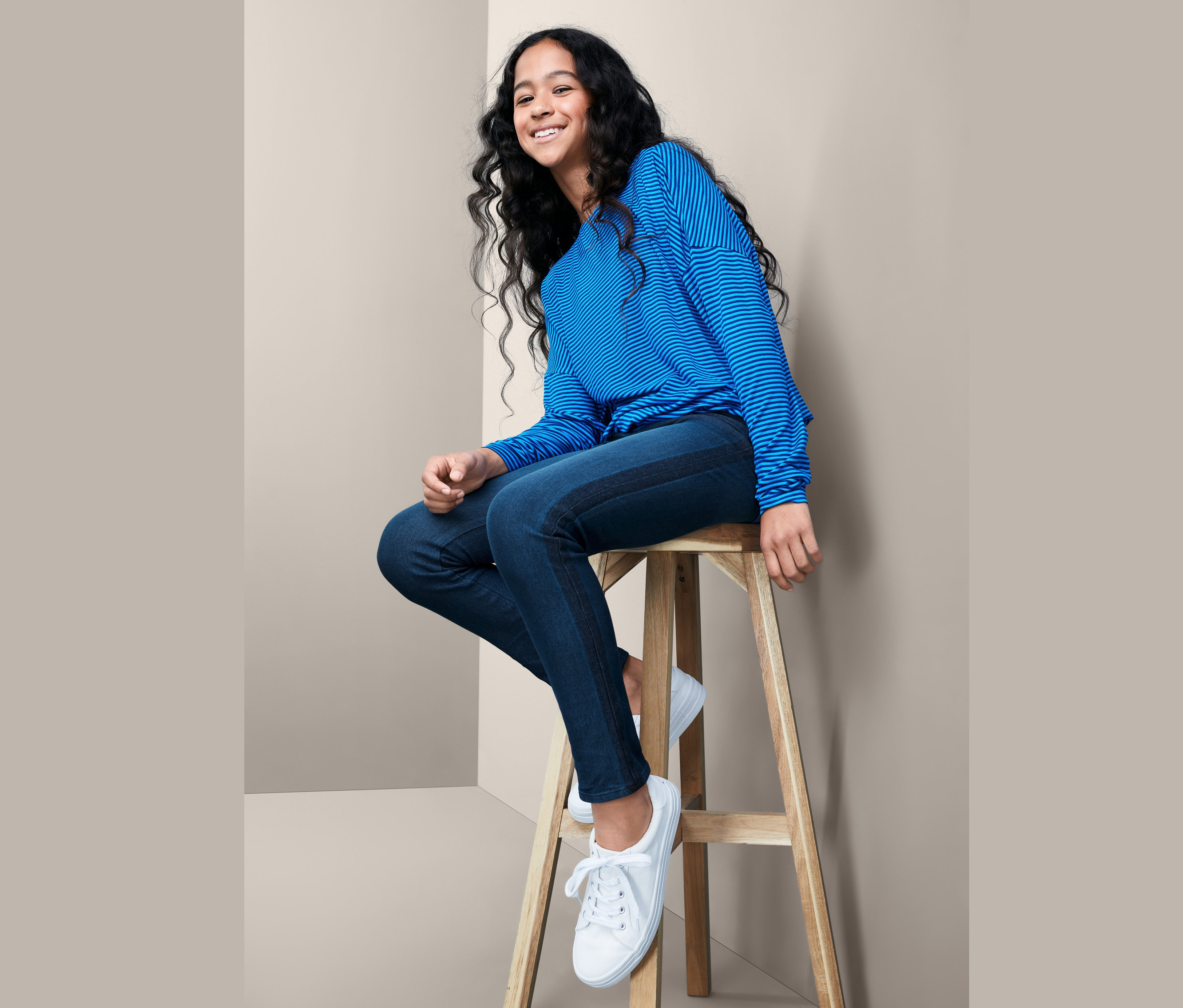 Ein Mädchen sitzt auf einem Holzhocker und trägt ein blau gestreiftes Oberteil und eine Stretch-Jeans.