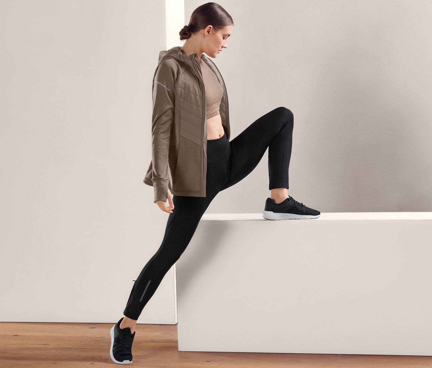 Eine Frau dehnt sich in schwarzen Performance-Lauftights, einem Sport-BH und einer braunen Jacke.
