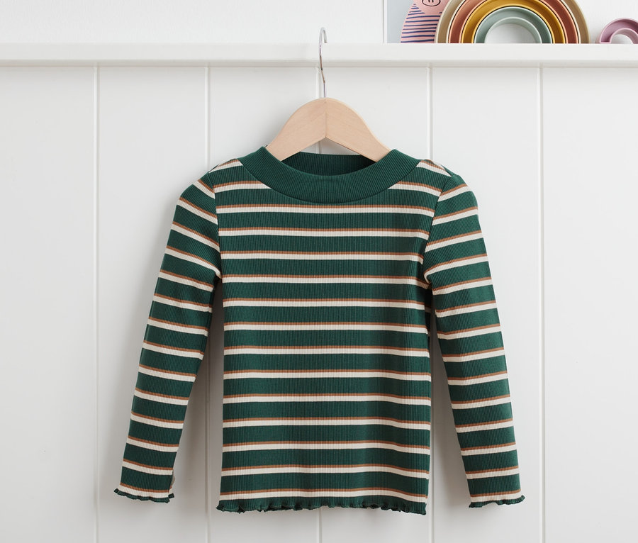 Ein grün geringeltes Rippshirt hängt auf einem Bügel vor einer weißen Wand.