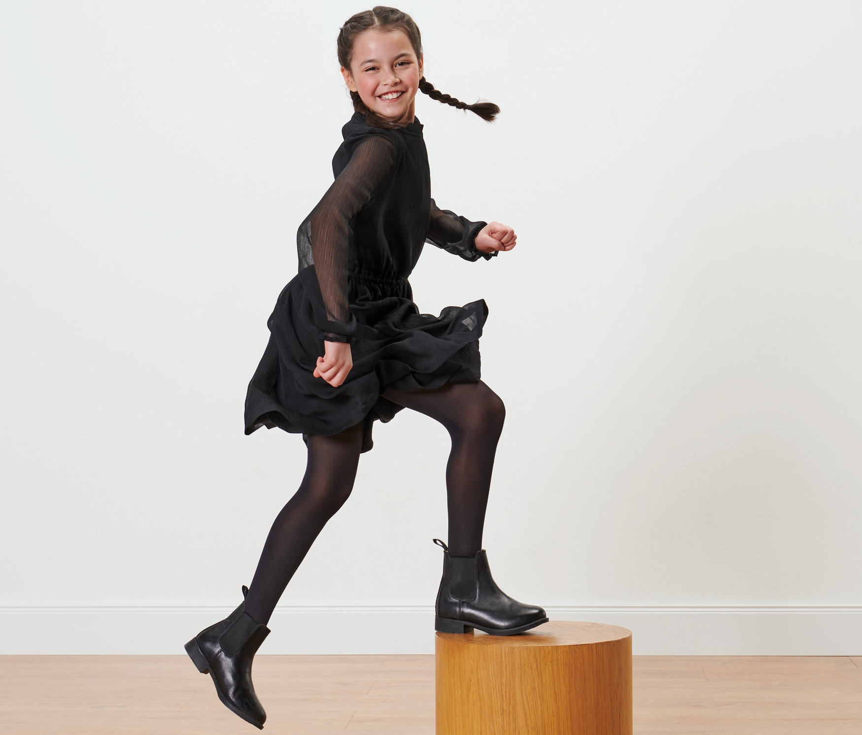 Lächelndes Mädchen mit Zöpfen in schwarzem Kleid und schwarzen 2 Kinder-Nylon-Strumpfhosen springt von einem Holzsockel.